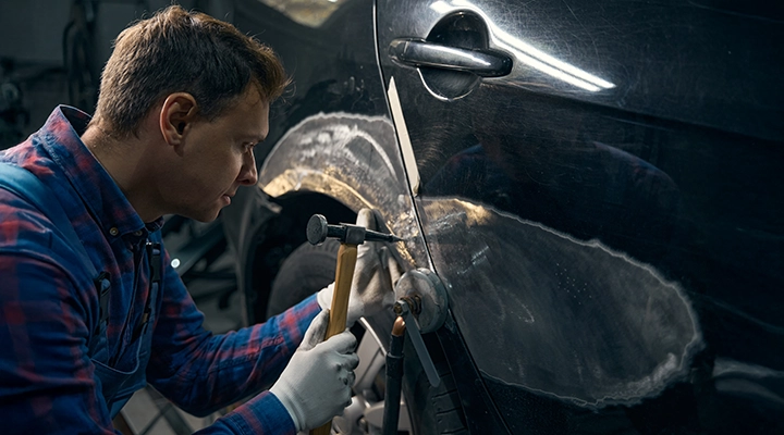 Auto body technician using a hammer and dolly to reshape and repair a damaged car panel in a workshop.