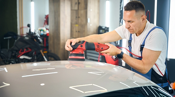 Auto detailing technician using a dual-action polisher to remove surface scratches and restore shine on a car hood in a workshop.