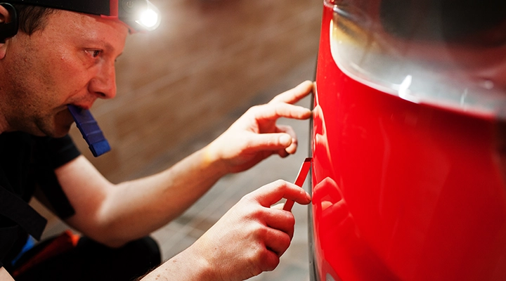 Technician closely inspecting and repairing a small dent on a red car panel using precision tools and reflection lighting.