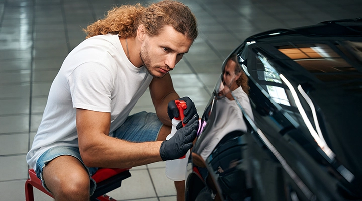 Technician carefully repairing minor scratches on a car panel using detailing tools and cleaning solution in an auto workshop.