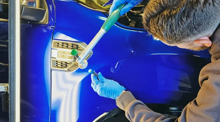 Technician performing paintless dent repair on a blue car panel using specialized tools to remove a small dent.