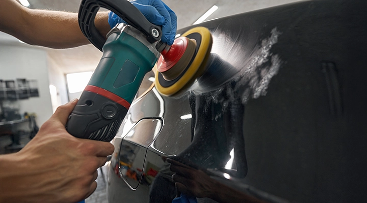 Technician polishing a car surface with a power buffer to remove scratches and restore shine in an auto body workshop.