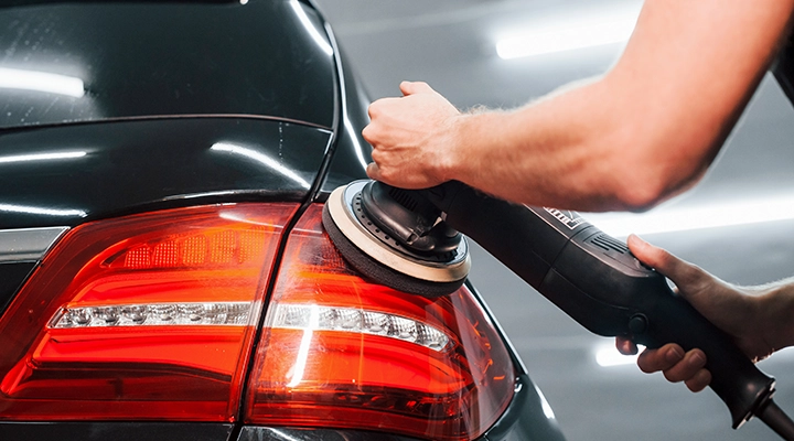 Technician using a power buffer to polish a car’s rear panel and taillight area, restoring gloss and removing surface scratches.