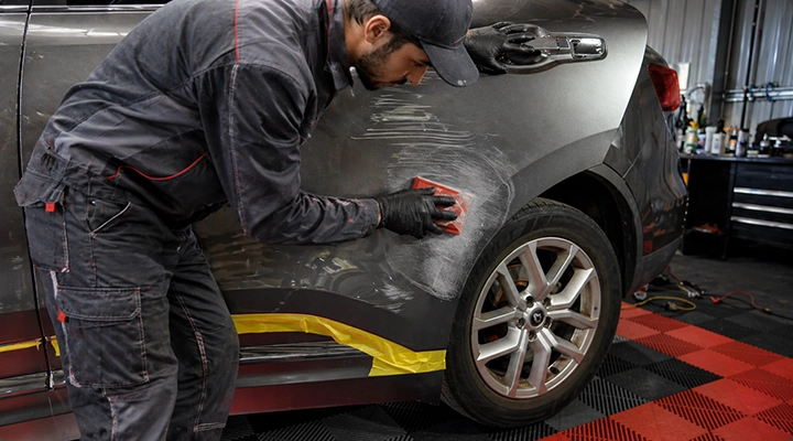 Auto body technician in protective workwear sanding and repairing scratches on a car’s rear panel in a professional workshop.
